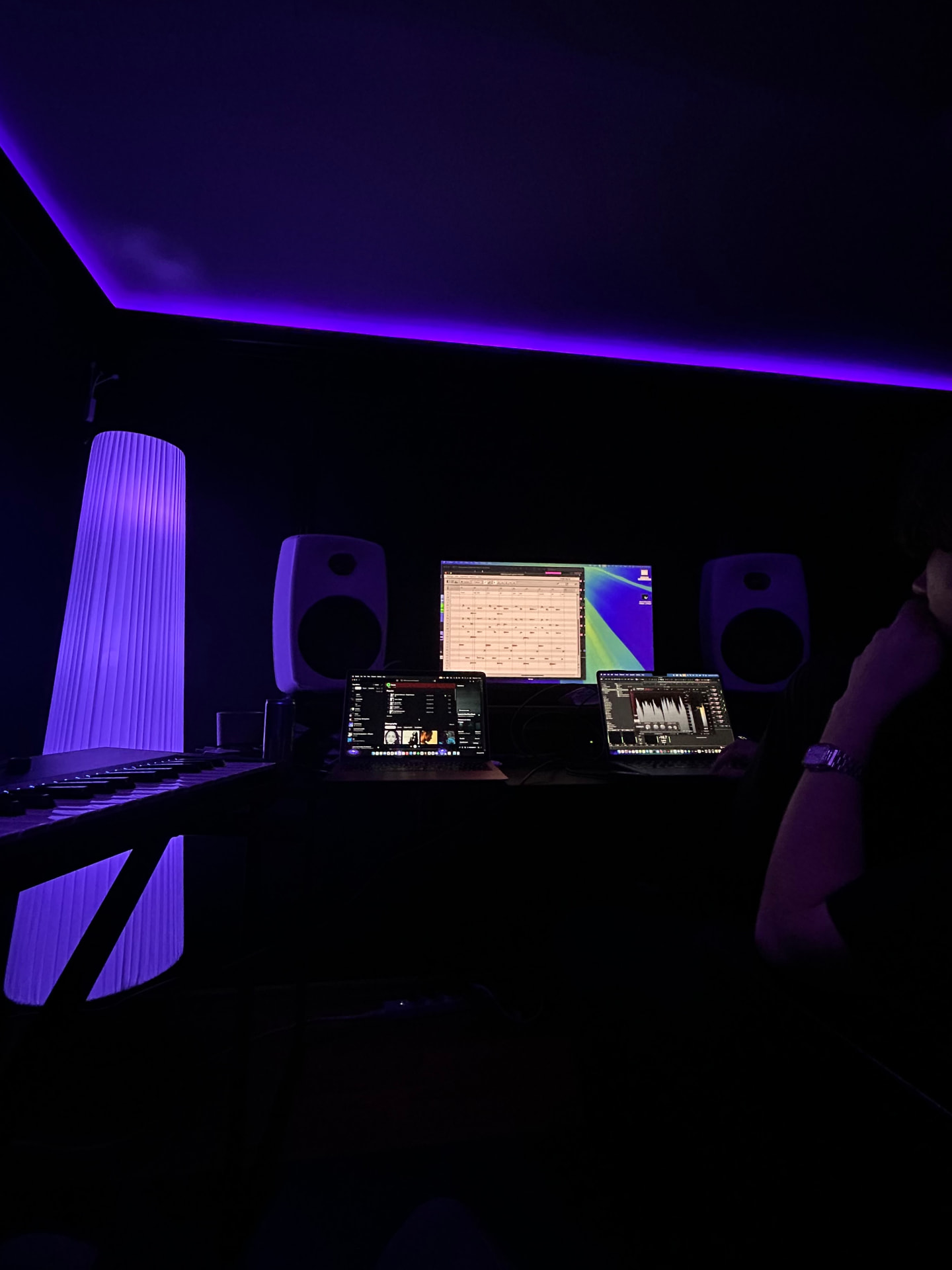 A purple-lit studio with monitors, a glowing floor lamp and laptops open on a DAW