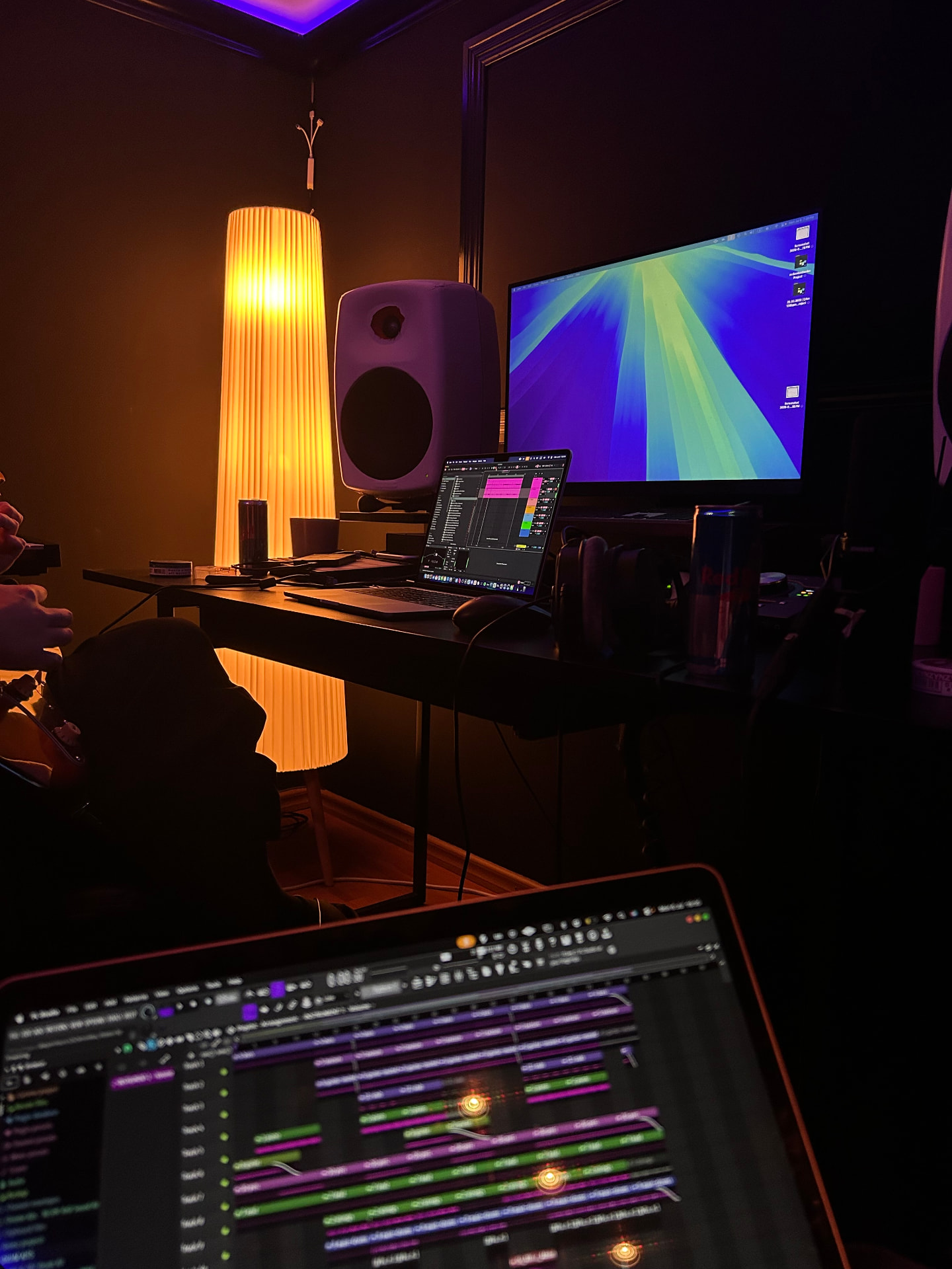 A warm lamp glowing over a studio desk with a monitor, speaker and laptop